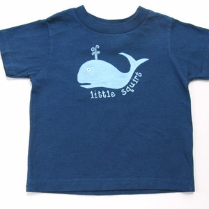 May include: A blue short-sleeved t-shirt with a light blue whale graphic and the text "little squirt" on the front.