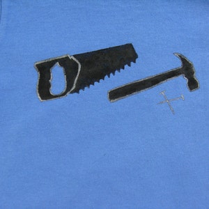 Pode incluir: Camiseta azul claro com uma serra preta, um martelo e dois pregos cruzados. As ferramentas t&ecirc;m uma contorno prateado. O design est&aacute; centrado na camiseta, sugerindo um tema relacionado &agrave; carpintaria ou constru&ccedil;&atilde;o.