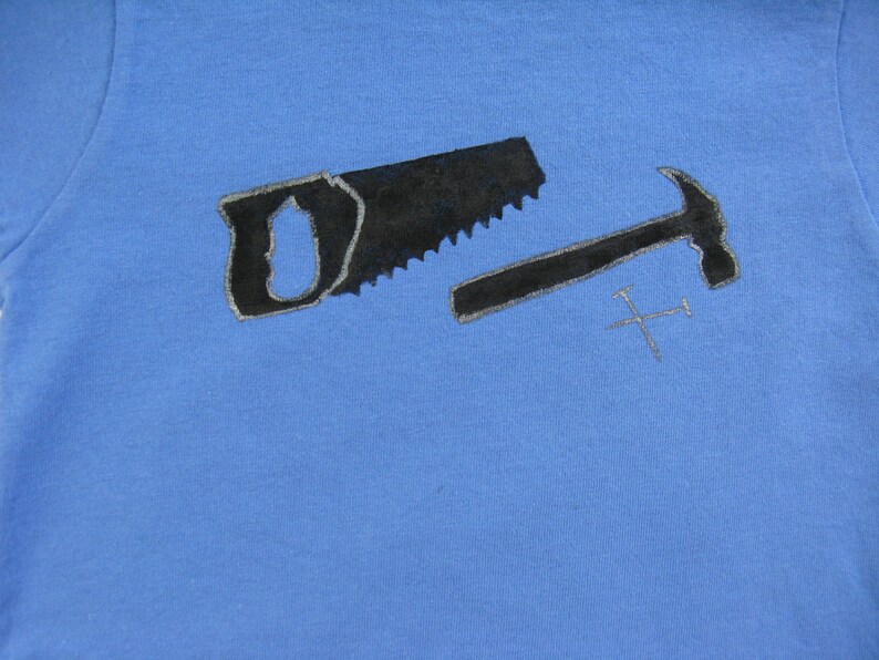Pode incluir: Camiseta azul claro com uma serra preta, um martelo e dois pregos cruzados. As ferramentas t&ecirc;m uma contorno prateado. O design est&aacute; centrado na camiseta, sugerindo um tema relacionado &agrave; carpintaria ou constru&ccedil;&atilde;o.