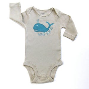 May include: A beige long-sleeved baby onesie with a blue whale graphic and the text "little squirt".