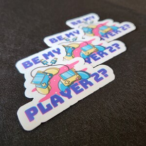 May include: Four white stickers with a pink and blue design featuring a video game controller and the text "Be My Player 2?"