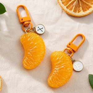 May include: Two orange-colored keychain charms shaped like citrus segments. Each charm features a bright orange lobster clasp and a small, round white pendant with a black letter, one displaying an X and the other displaying an E.