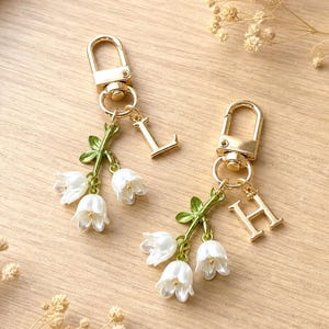 May include: Two gold-toned metal keychains featuring white lily of the valley flower charms with green stems and leaves. Each keychain includes a lobster clasp and a single hanging letter charm, specifically the letters L and H in a serif font.