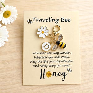 May include: A gold-toned metal charm featuring a white daisy, a circular pendant with the letter D, and a bee with crystal-encrusted wings. The charm is attached to a display card with the text Traveling Bee and a poem about journeys.