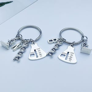 Puede incluir: Dos llaveros plateados con colgantes de temática científica. Cada llavero tiene un anillo, un matraz, una cadena de ADN, un microscopio y un dije de letra. Los matraces tienen una escala de 100 ml. El fondo es de color claro.