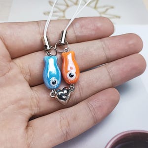 Può includere: Un paio di ciondoli per telefono abbinati. Uno è blu, l'altro arancione, entrambi con un design a occhio dei cartoni animati. Sono collegati da un ciondolo a cuore argentato e cordini bianchi.