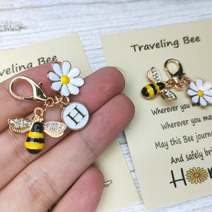 Könnte beinhalten: Goldfarbener Charm mit einem Anhänger in Form einer Biene, einer Gänseblümchenblüte und dem Buchstaben "H". Die Biene hat schwarz-gelbe Streifen und Strassflügel. Das Gänseblümchen ist weiß mit gelber Mitte. Der Charm liegt neben einer Karte mit "Traveling Bee".