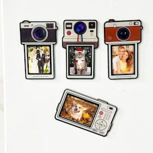 May include: Camera-shaped photo frames with pictures of a wedding, a cat in a Santa hat, a couple, and a dog. The frames are black and white with a retro design. The dog photo frame has buttons and a screen.