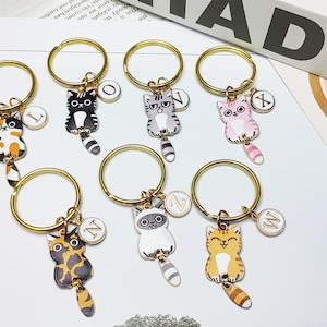 May include: A collection of cat-shaped keychains with gold-colored rings. Each keychain features a different cat design in various colors, including orange, black, gray, and pink. Each cat has a dangling tail and a small, round charm with a letter.