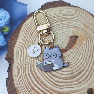 K&ouml;nnte beinhalten: Ein goldfarbener Schl&uuml;sselanh&auml;nger mit einem Katzenanh&auml;nger, der eine Brille tr&auml;gt und ein Buch liest. Der Anh&auml;nger ist grau und wei&szlig; mit schwarzen Details. Ein wei&szlig;er runder Anh&auml;nger mit dem Buchstaben "V" ist ebenfalls angebracht. Der Schl&uuml;sselanh&auml;nger befindet sich auf einer Holzoberfl&auml;che.
