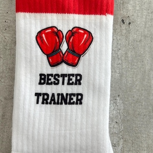 Puede incluir: Un calcetín blanco de caña media con un puño a rayas negras y rojas en la parte superior. El calcetín muestra un gráfico de dos guantes de boxeo rojos sobre el texto negro BESTER TRAINER en una fuente negrita y sin remates sobre la tela acanalada.