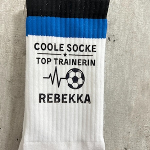 Puede incluir: Un calcetín blanco con un puño a rayas negras y azules. El calcetín presenta el texto negro COOLE SOCKE TOP TRAINERIN REBEKKA, acompañado de un gráfico de una línea de latido del corazón y un balón de fútbol impreso en la parte frontal.