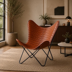 May include: A brown leather butterfly chair with a black metal frame. The chair has a distinctive, curved shape and is placed in a living room setting. The leather has a textured appearance.