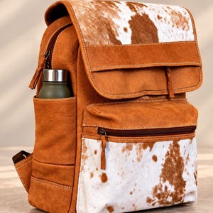 Könnte beinhalten: Ein beigefarbener Lederrucksack mit einer Klappe und einer Vordertasche aus weiß-braun geflecktem Kuhfell. Die Tasche verfügt über ein Seitenfach mit einer kleinen olivgrünen Trinkflasche mit silbernem Verschluss, braunen Reißverschlüssen und weißen Nähten.
