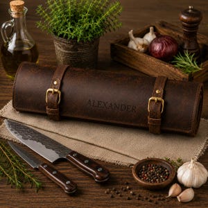Può includere: Un rotolo portacoltelli in pelle marrone scuro con due fibbie in ottone e il nome ALEXANDER impresso sulla parte anteriore. Due coltelli da cucina con manici in legno scuro e lame in metallo martellato sono posti davanti al rotolo su un panno beige.