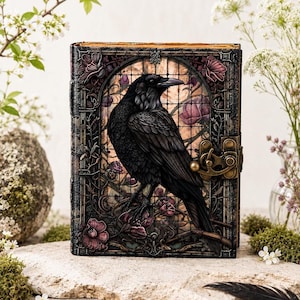 May include: A rectangular journal featuring a black raven illustration centered within an arched frame. The cover displays a stained glass pattern with purple flowers and a metallic brass-colored latch closure on the right side edge.