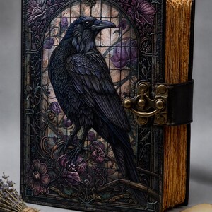 May include: A hardcover journal featuring a black raven illustration set against a stained glass background with purple flowers. The book has a dark leather-like cover, a brass metal latch closure, and thick, textured, light brown paper pages.