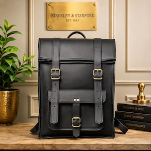 Puede incluir: Una mochila negra con silueta rectangular, asa superior y dos correas verticales con hebillas de tono latón. Un bolsillo frontal está asegurado con una correa central y una hebilla. El diseño presenta superficies lisas y costuras visibles.
