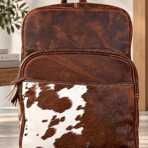May include: A brown leather backpack featuring a top handle and a front pocket with a cowhide panel. The backpack has a distressed, dark brown finish on the upper section and a white and brown spotted cowhide pattern on the lower front pocket.