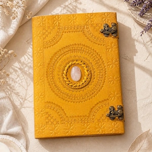 Puede incluir: Un diario rectangular de color amarillo mostaza con un patrón floral y geométrico en relieve en la cubierta. Una piedra lisa de color rosa claro y forma ovalada está centrada en un borde trenzado. Dos cierres de metal color latón antiguo.