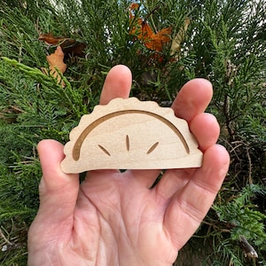 May include: A wooden cutout of a half-moon shape with a scalloped edge. The cutout is light brown and has a simple design of lines radiating from the center.