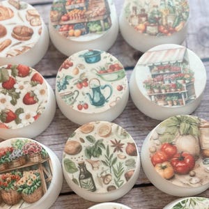 May include: A collection of white, round objects with various food-themed images on top. Designs include bread, strawberries, a fruit stand, flowers, and tomatoes. The objects are arranged on a wooden surface.