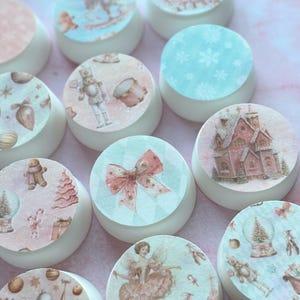 May include: A collection of white, round objects with decorative, circular tops. The tops feature various watercolor illustrations, including a nutcracker, a gingerbread man, a snow globe, and a pink bow. The overall theme is festive and holiday-oriented.