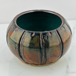May include: A small, round ceramic bowl with a dark green interior and a textured exterior. The bowl has a brown, black, and green glaze with a metallic sheen.