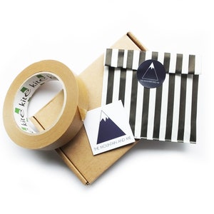 May include: A brown cardboard box with a roll of brown packing tape and a white sticker with a blue mountain design and the text "The Mountain and Me". A black and white striped paper bag with a black circle sticker with a white mountain design and the text "The Mountain and Me" is on top of the box.