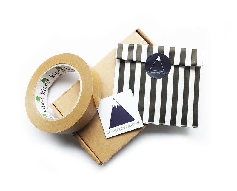 May include: A brown cardboard box with a roll of brown packing tape and a white sticker with a blue mountain design and the text "The Mountain and Me". A black and white striped paper bag with a black circle sticker with a white mountain design and the text "The Mountain and Me" is on top of the box.