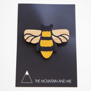 May include: A wooden bee brooch painted in yellow and black with a natural wood finish. The bee is on a black background with a white triangle logo that says "The Mountain and Me".