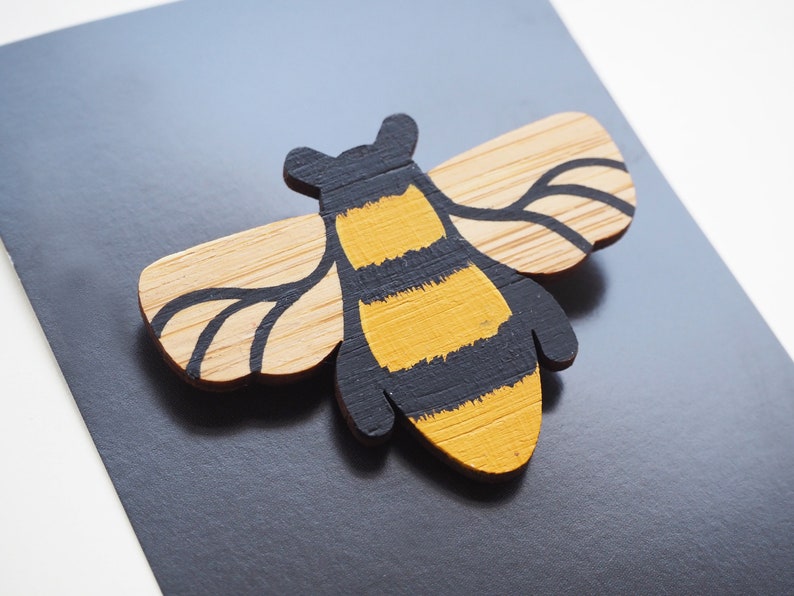 May include: A wooden bee brooch painted with yellow and black stripes. The bee has wings made of natural wood with black painted details.