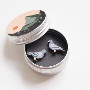 May include: A small, round, silver tin with a lid featuring a mountain scene and the text "HOME". Inside, two wooden pigeon-shaped pins rest on a black surface. The pins are gray, blue, and brown.