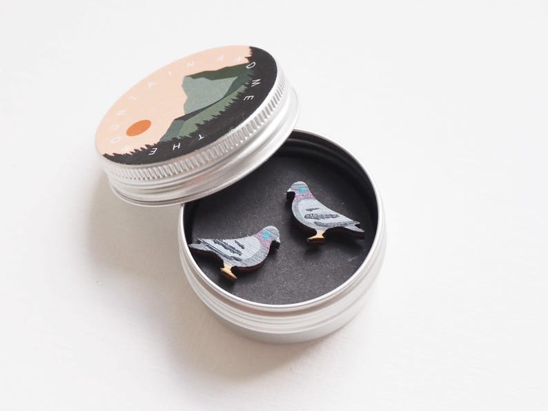 May include: A small, round, silver tin with a lid featuring a mountain scene and the text "HOME". Inside, two wooden pigeon-shaped pins rest on a black surface. The pins are gray, blue, and brown.