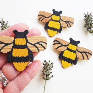 May include: Three wooden bee brooches painted in black and yellow with brown wings. The bees are on a white background.