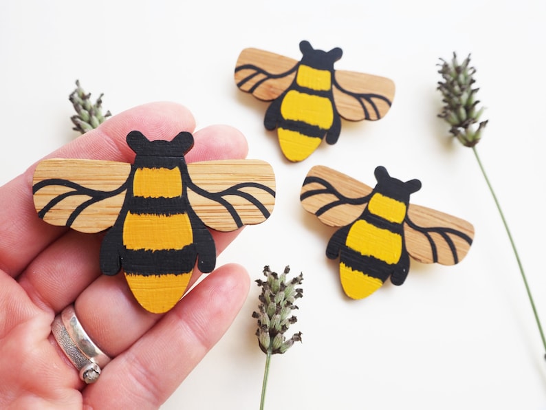 May include: Three wooden bee brooches painted in black and yellow with brown wings. The bees are on a white background.