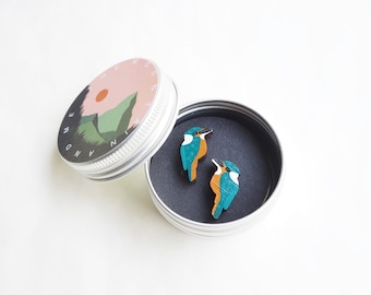 May include: Two small wooden earrings shaped like blue and orange kingfishers. The earrings are in a small round tin with a black interior. The tin has a label that says "The Mountain and Me".