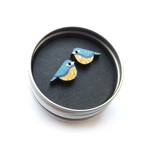 以下が含まれることがあります： 青と黄色の鳥が描かれた木製イヤリングのペア。イヤリングは丸いブリキの容器に入っています。