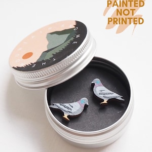 May include: A small, open silver tin containing two hand-painted wooden pigeon earrings. The earrings are gray, blue, and brown. The tin lid has a mountain scene with the text "MOUNTAIN HOME". The text "HAND PAINTED NOT PRINTED" is also visible.