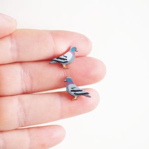 May include: Two miniature pigeon-shaped earrings. Each earring features a gray body with black wing markings, a purple head, and yellow feet. The earrings are small, detailed, and appear to be made of wood or a similar material. The earrings are on a white background.