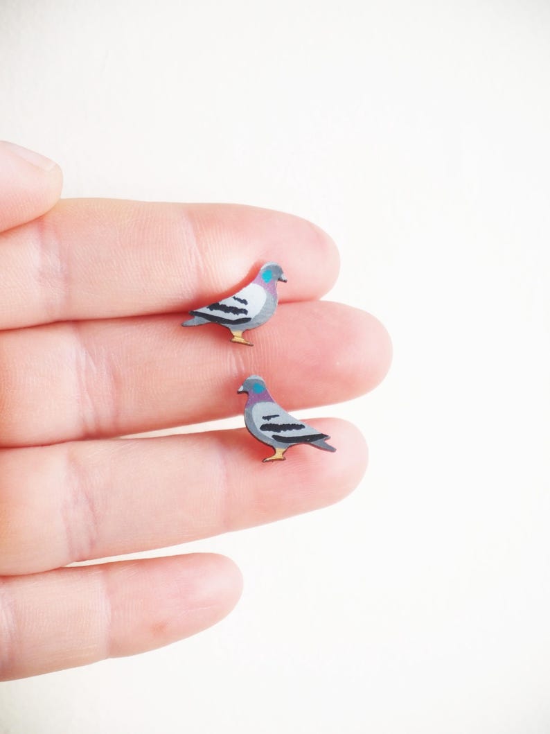 May include: Two miniature pigeon-shaped earrings. Each earring features a gray body with black wing markings, a purple head, and yellow feet. The earrings are small, detailed, and appear to be made of wood or a similar material. The earrings are on a white background.