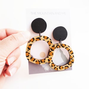 May include: A pair of leopard print wooden earrings with black circle accents. The earrings are hanging from a card that says "The Mountain and Me."