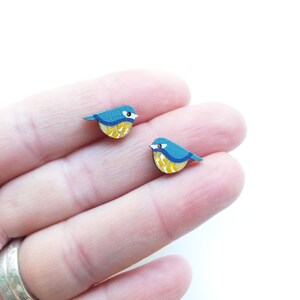 以下が含まれることがあります： 青いシジュウカラの形をした木製のイヤリングのペア。鳥は黄色、青、白のディテールで描かれています。