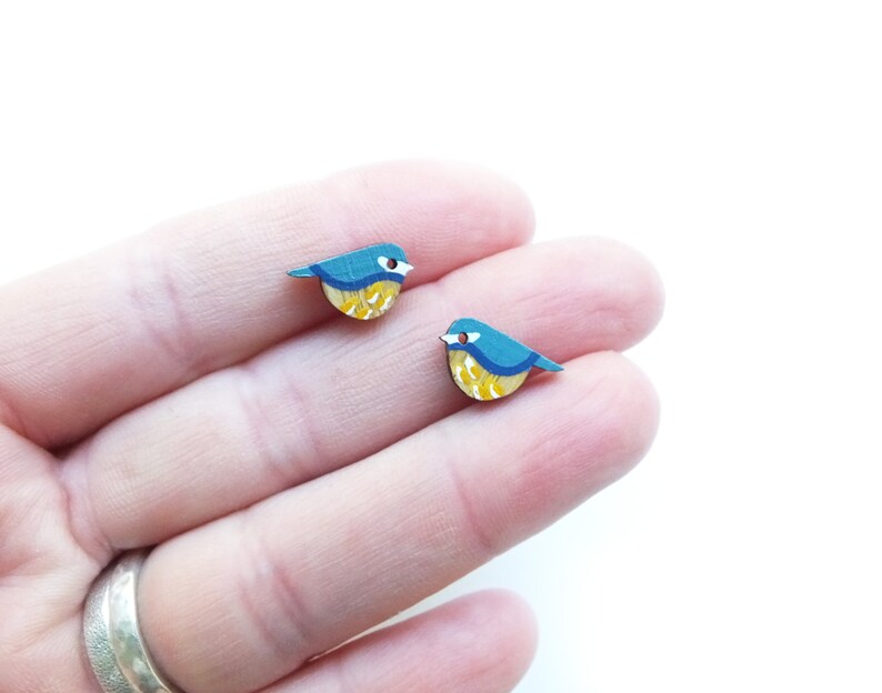 以下が含まれることがあります： 青いシジュウカラの形をした木製のイヤリングのペア。鳥は黄色、青、白のディテールで描かれています。