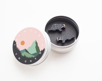 May include: A pair of black badger earrings in a small round tin with a white lid. The lid has a design of a mountain range with the text "The Mountain and Me".