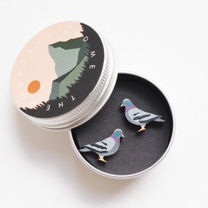 May include: A small, open, silver tin containing two pigeon-shaped pins. The pins are gray, black, and purple with yellow feet. The tin lid features a landscape design with the text "THE MOUNTAIN AND ME" in a circular pattern.