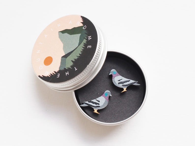 May include: A small, open, silver tin containing two pigeon-shaped pins. The pins are gray, black, and purple with yellow feet. The tin lid features a landscape design with the text "THE MOUNTAIN AND ME" in a circular pattern.