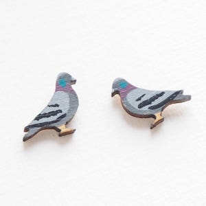 May include: Two wooden pigeon-shaped ornaments. The birds are painted gray with black wing details, a teal head spot, and a purple neck. The feet are a light brown color. The ornaments are likely for decoration or crafting.
