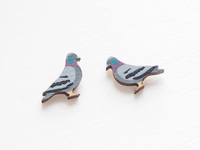 May include: Two wooden pigeon-shaped ornaments. The birds are painted gray with black wing details, a teal head spot, and a purple neck. The feet are a light brown color. The ornaments are likely for decoration or crafting.
