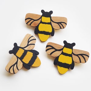 May include: Three wooden bee ornaments painted with black and yellow stripes. The bees have wings and are made of natural wood.